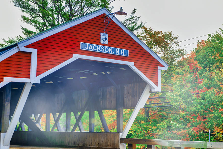a view of a wooden and red covered bridge with the words "Jackson, NH" on the front of the bridge.
