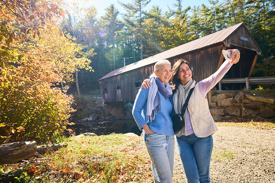 two women taking a picture in front of a covered bridge in fall.