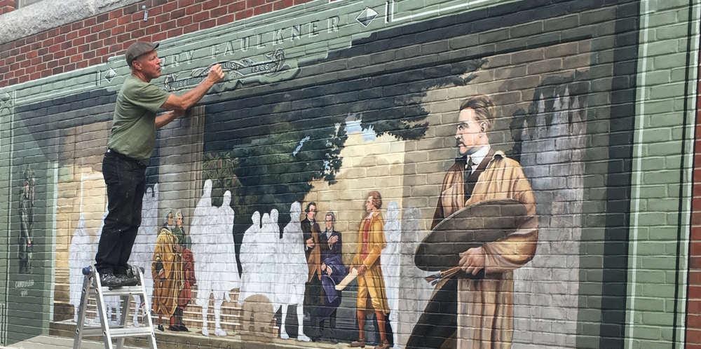 a man painting a mural standing on a step stool outdoors.