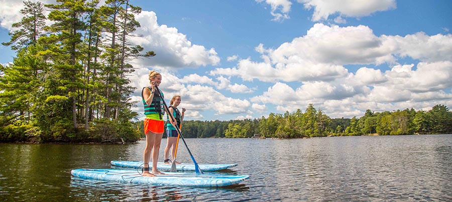 Two women on stand up paddleboards in a lake surrounded by green trees.