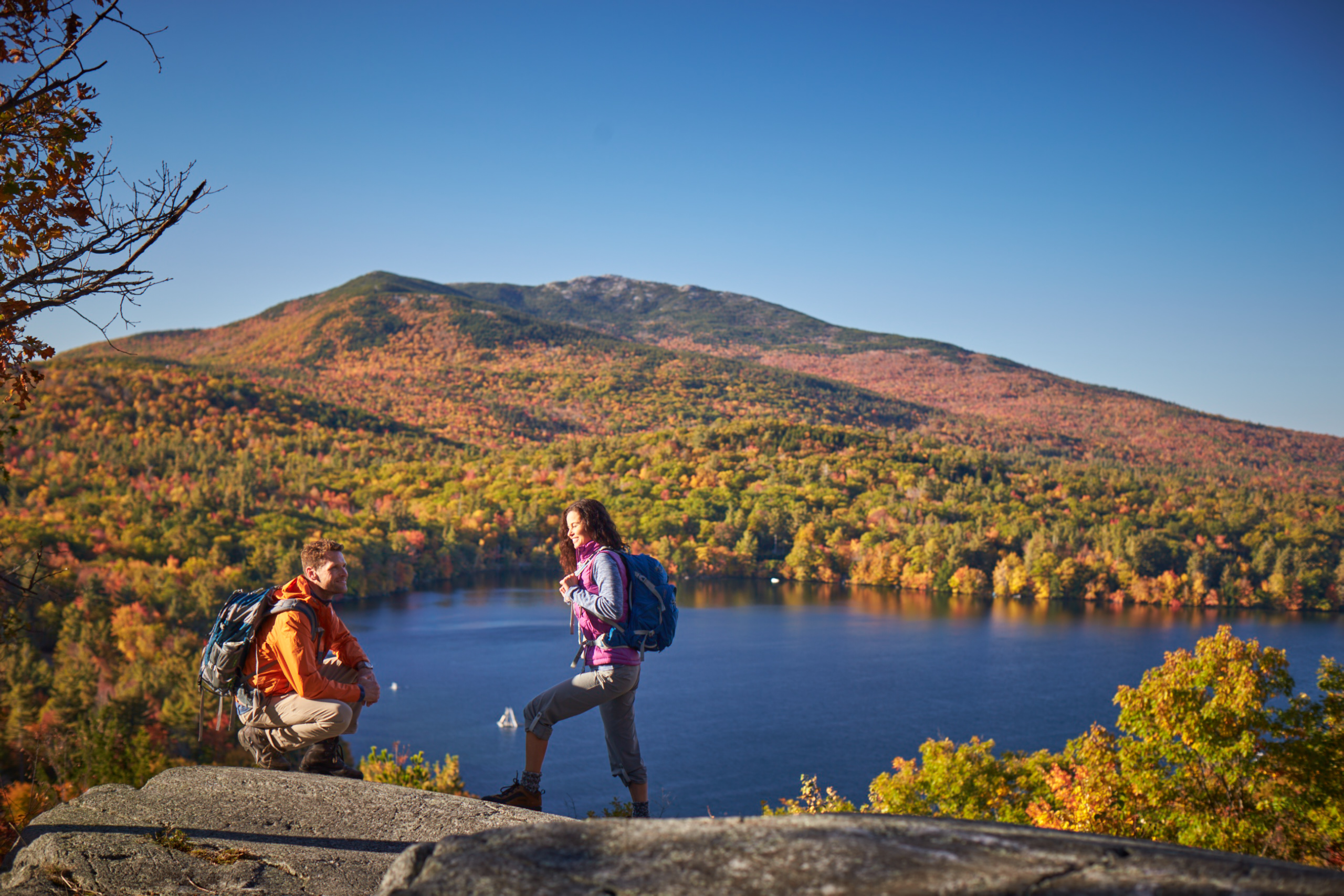 a couple hiking in the fall with views of a hillside covered in foliage and a lake.