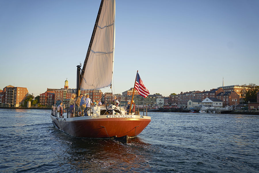 a gundalow boat with an American flag on the front sailing in front of the Portsmouth shoreline.