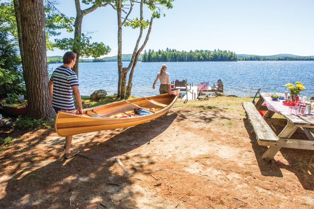 A woman and man carrying a canoe towards a lake, next to a picnic table set up with food.